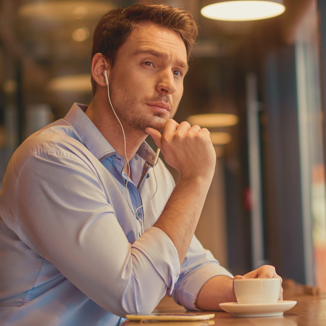 A man in a light blue collared shirt with earbuds in listens thoughtfully while resting his chin on his hand next to a coffee cup and phone.